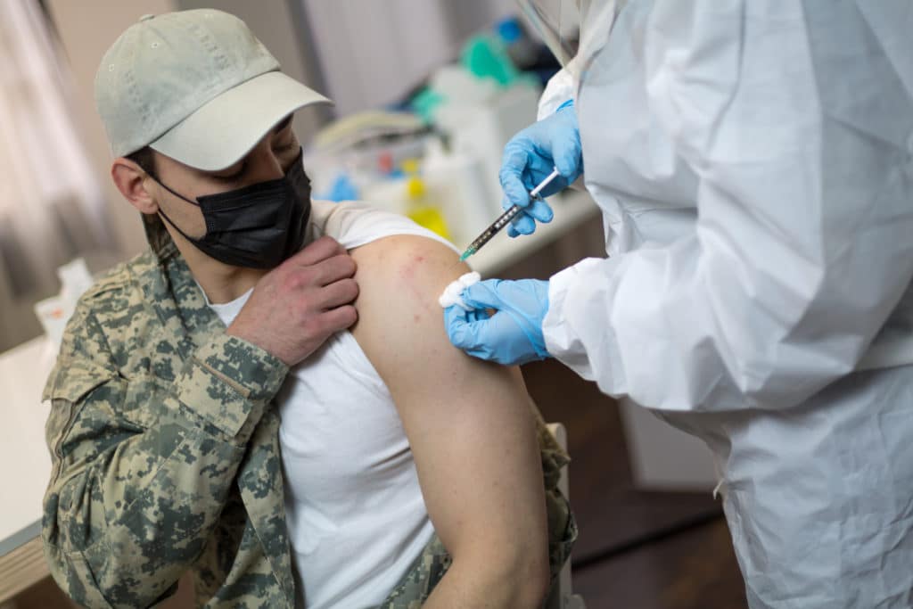Young female doctor in protective suit injecting COVID-19 vaccine in arm of a young military soldier for coronavirus immunization.
