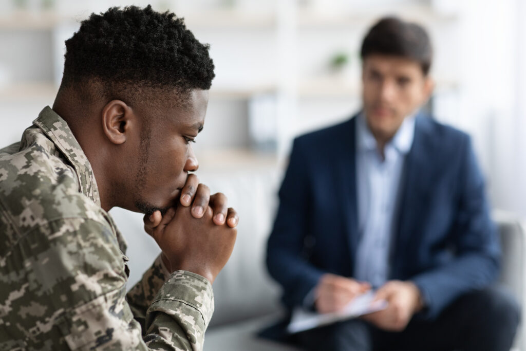 Pensive black man in camouflage uniform soldier having conversation with social worker, selective focus on african american military guy suffering from PTSD after returning home, blurred background