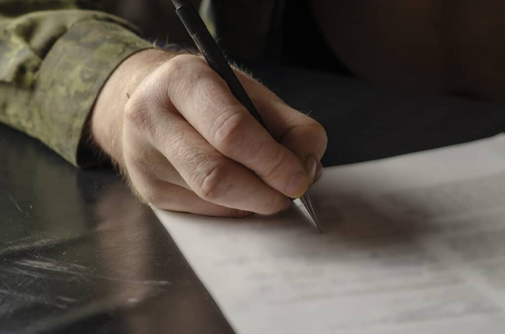 A middle-aged adult man in a green camouflage uniform signs a document. A hand makes a signature with a black pen. Indoors. Selective Focus. Low key. Close-up.